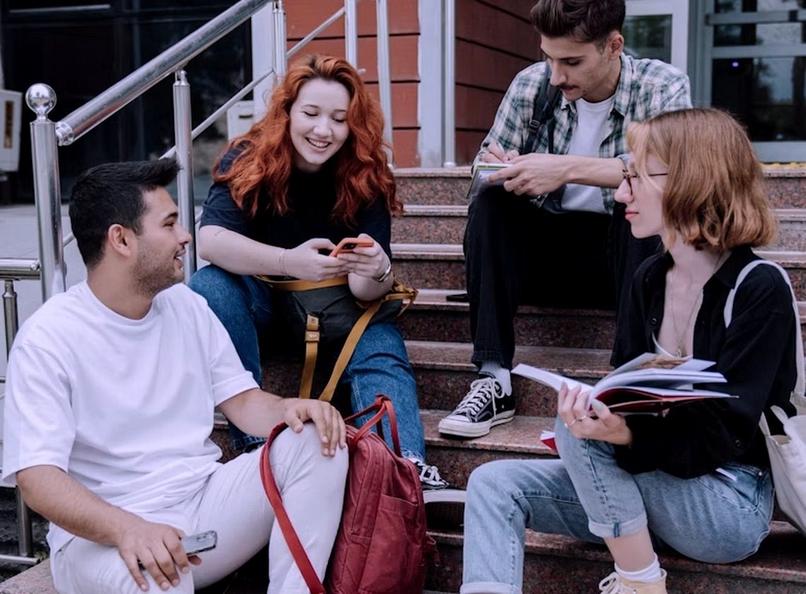 Students sitting on campus steps, talking and checking a phone